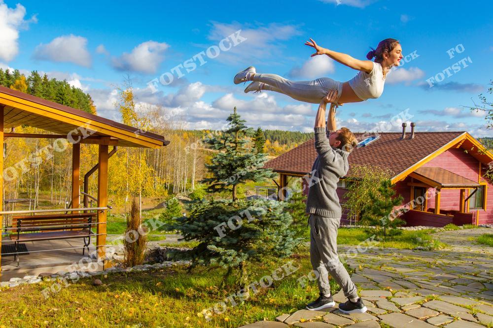 На фотографии мужчина поднимает девушку высоко над головой на фоне красивого загородного дома и осеннего пейзажа. Яркие краски природы, солнечный день и гармония создают атмосферу радости и свободы. Снимок символизирует активный образ жизни, любовь и совместный отдых на природе.